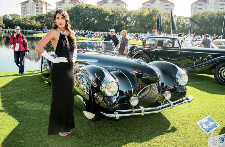 A guest in a black evening gown and white gloves beside a black prewar car on the show field at the Amelia Island Concours d’Elegance, an engagement in the season calendar of The Mansa Court