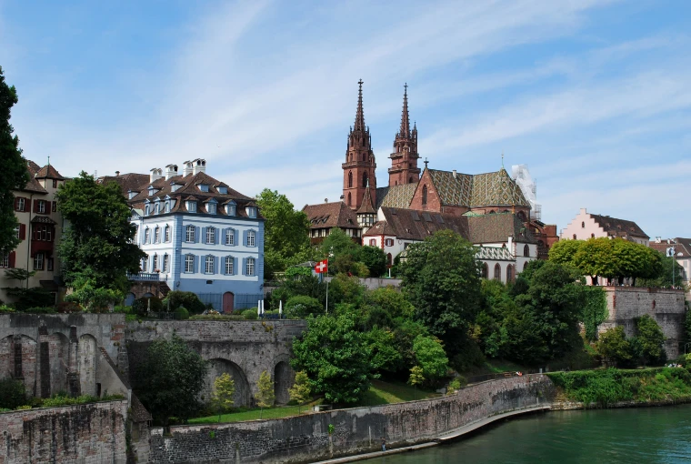 The red sandstone twin spires and patterned tile roof of Basel Minster rise above green trees, old houses and the Pfalz terrace wall on the bank of the Rhine under a summer sky (2015). Basel, Switzerland, shown for Art Basel in the season calendar of The Mansa Court