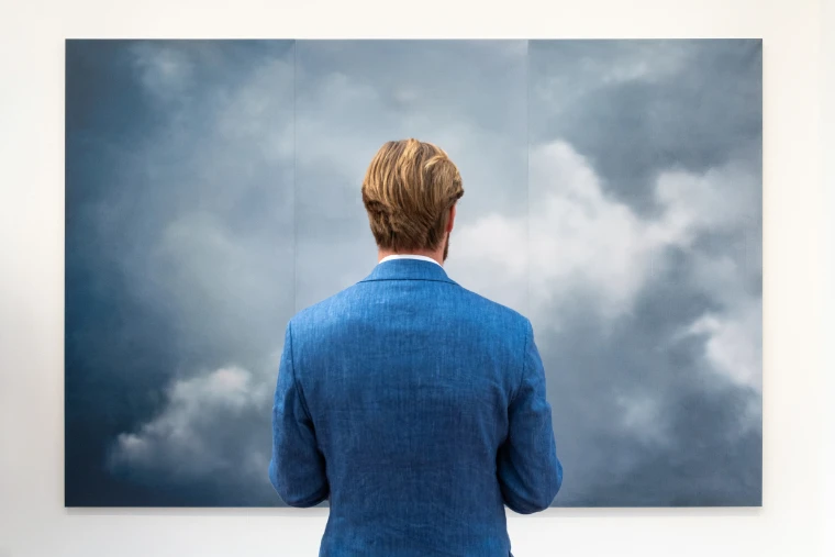 A man in a blue jacket stands before a large painting of grey clouds on a white gallery wall (2025).