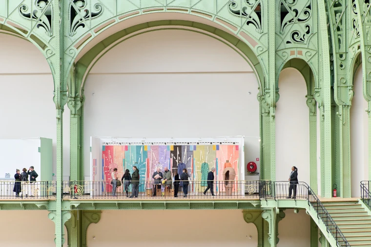 Visitors walk along the balcony of the Grand Palais past a colourful booth, framed by pale green ironwork arches (2025).