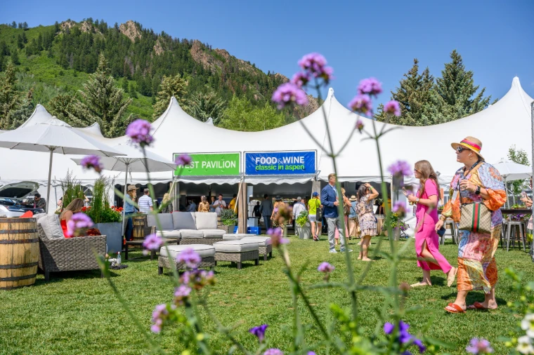 Guests in summer dress crossing the lawn and lounge outside the tasting pavilion, wildflowers in the foreground and the mountain behind, at the Aspen Food and Wine Classic, the June festival in the season calendar of The Mansa Court