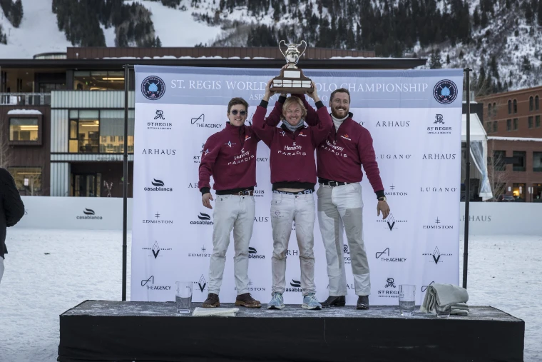 Three players in maroon jackets raise a silver trophy on the prize-giving stage, with the snowy ski slopes of Aspen Mountain behind (2024).