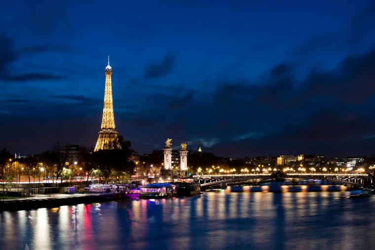 The Eiffel Tower lit gold against a deep blue night sky, with the Pont Alexandre III and the lights of the quays reflected in the Seine (2017).