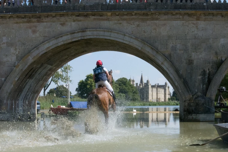 A horse and rider splash through the lake beneath a stone bridge arch, with Burghley House framed beyond (2018).