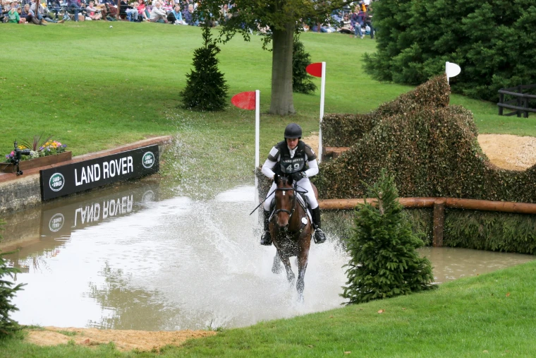 A horse and rider canter out of a water complex in a spray of water beside a Land Rover sign, with spectators on the bank above (2011).