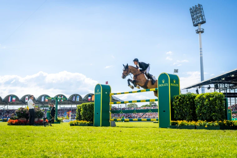 A horse and rider sailing over the green and yellow Rolex oxer beneath a summer sky before the stands at CHIO Aachen, a July engagement in the season calendar of The Mansa Court