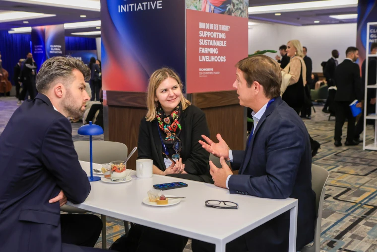 Three delegates with name badges talk across a small white table with coffee and pastries, beside a sign reading “How do we get it right? Supporting sustainable farming livelihoods” (2026).