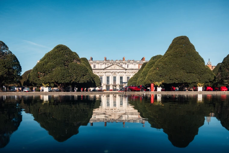 The east front of Hampton Court Palace between clipped yew cones, with a line of concours cars along the far bank of the Long Water and the whole scene mirrored in the still water (2026).