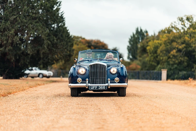 A dark blue Bentley drophead with its hood down drives towards the camera along a gravel avenue, a driver in a panama hat at the wheel (2026).
