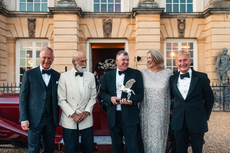 Five people in evening dress stand on the gravel before the palace’s stone doorway, one man holding a silver winged-horse trophy, with a dark red vintage car at the left edge (2026).