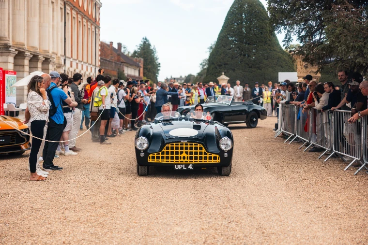 A black vintage sports car with a yellow grille drives down a gravel avenue between crowds behind barriers, the palace front on the left and a clipped yew ahead (2026).