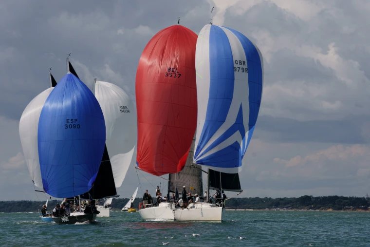 Racing yachts flying red, blue and white spinnakers run downwind together on grey-green water under a cloudy summer sky (2019).