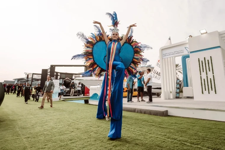 A stilt performer in feathered costume crossing the lawn between the stands at the Dubai International Boat Show, an engagement in the season calendar of The Mansa Court