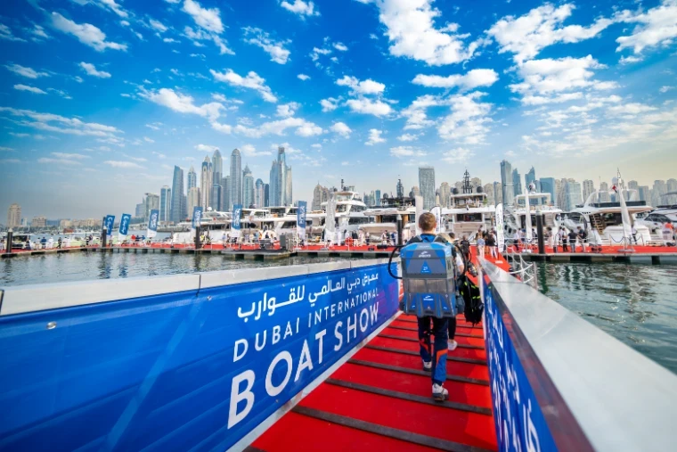 Visitors crossing onto the red-carpet pontoons past the show branding with yachts and the Dubai Marina skyline beyond at the Dubai International Boat Show, an engagement in the season calendar of The Mansa Court