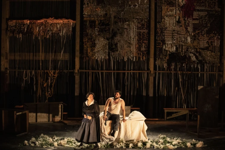 A performer in a long black dress stands beside another seated on a white-draped bed amid scattered flowers under warm light, before a tall backdrop of hanging textiles (2026).