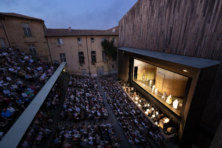 Seen from above at dusk, a packed open-air courtyard theatre faces a lit stage set against the pale stone walls of the Archbishop’s Palace (2025).
