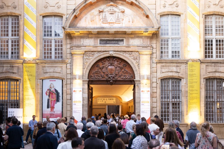 An evening crowd gathers before the carved doorway of the Archbishop’s Palace, its facade hung with yellow festival banners (2025).
