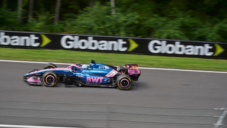 A pink and blue Alpine Formula 1 car speeds past a trackside advertising board with green trees behind (2026).