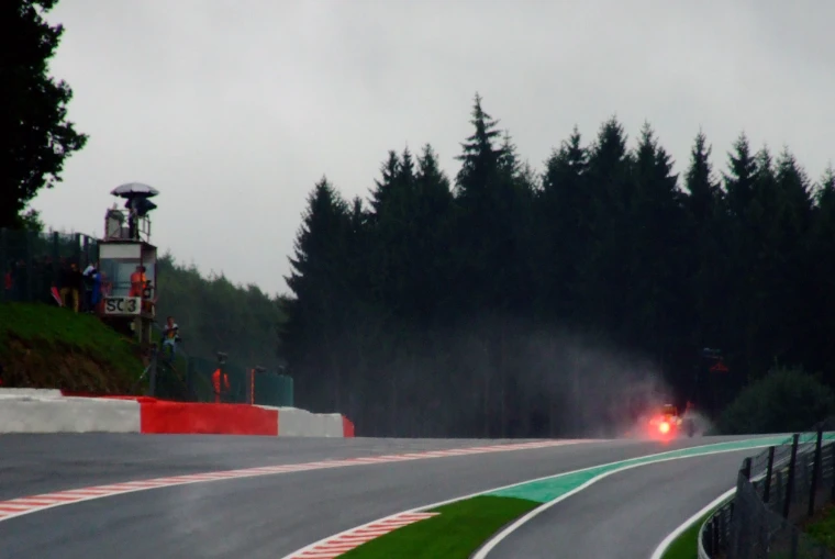 A Formula 1 car crests the Raidillon hill under a grey sky, its red rear light glowing in the spray beside a wall of dark pine trees (2010).