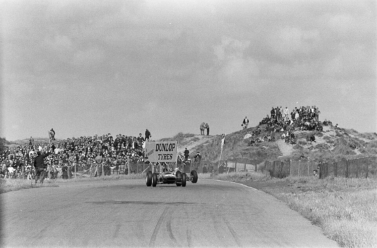 In black and white, two racing cars come towards the camera between sand dunes lined with spectators, beneath a Dunlop Tyres sign (1963).
