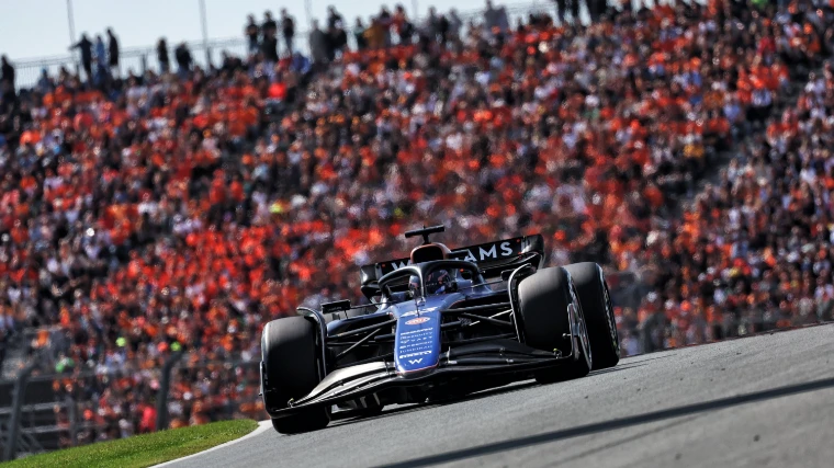 A dark-blue Williams Formula One car at speed through a corner, with a packed grandstand of fans in orange blurred behind it (2024).