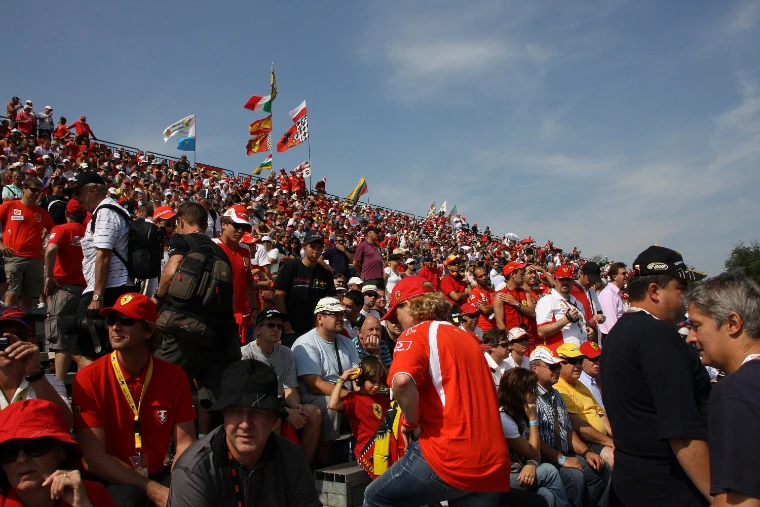 A steep grandstand packed with spectators in red Ferrari shirts and caps under a blue sky, with flags flying along its top (2010).