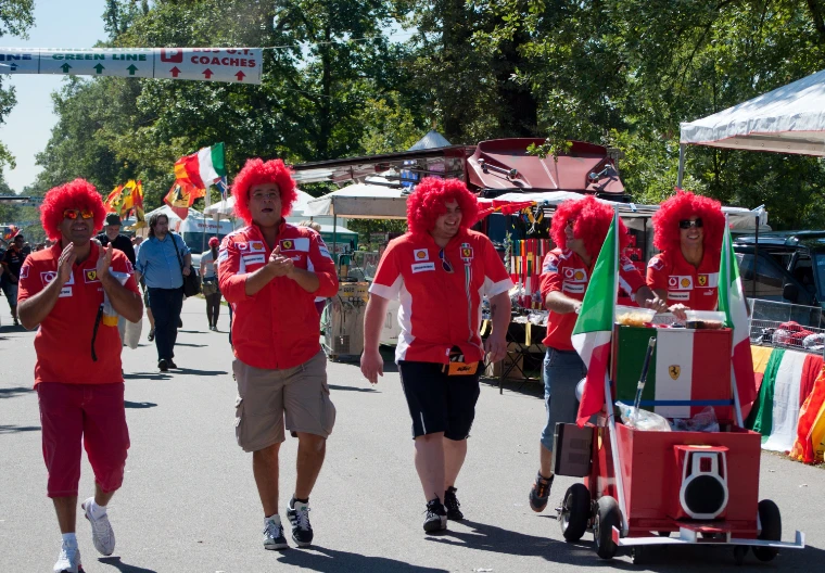 Five fans in red curly wigs and Ferrari shirts walk along a tree-lined road past merchandise stalls, one pushing a cart with Italian flags (2010).