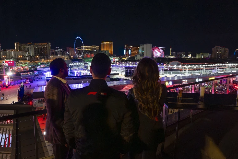 Three people seen from behind on a terrace look across the lit paddock to the Las Vegas skyline at night, with the glowing Sphere and the High Roller wheel on the horizon (2025).