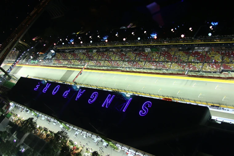 Looking down at night on the floodlit pit straight and packed grandstand, with SINGAPORE lit in purple letters on the roof of the pit building (2013).