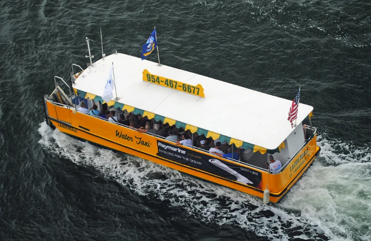 A yellow water taxi full of passengers under its striped awning cutting a white wake across dark water, seen from above (2021).