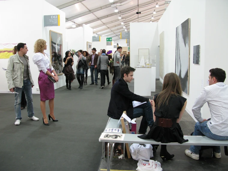 Three people talk on a bench beside the gallery booths at Frieze Art Fair while visitors walk the aisle behind them (2009).