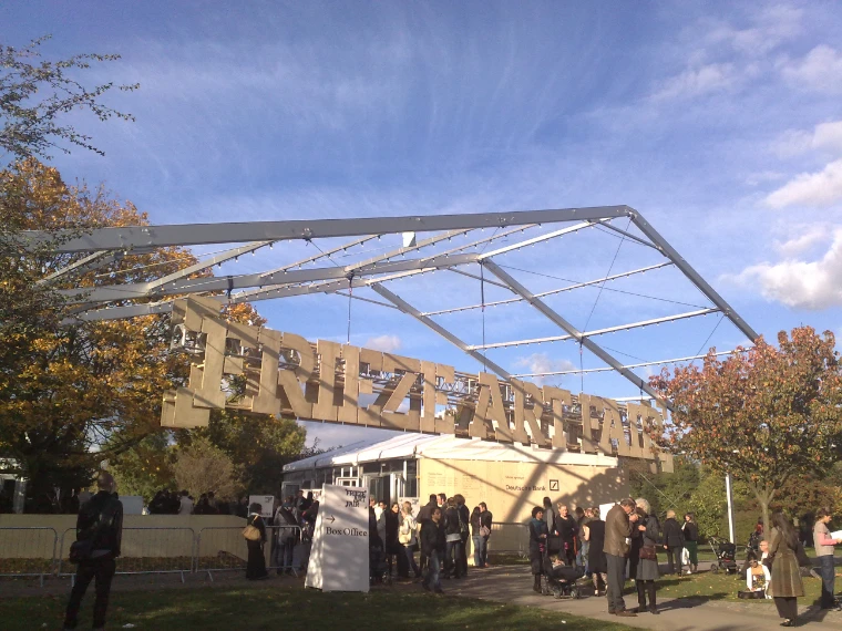 Visitors queue beneath a giant wooden “Frieze Art Fair” sign and steel canopy at the fair’s entrance, with the white tent and autumn trees under a blue sky (2008).