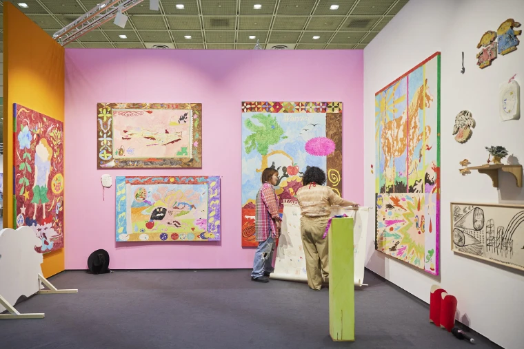 Two visitors unroll a long painted scroll in a gallery booth with a pink wall hung with brightly coloured painted banners and small wall pieces (2023).