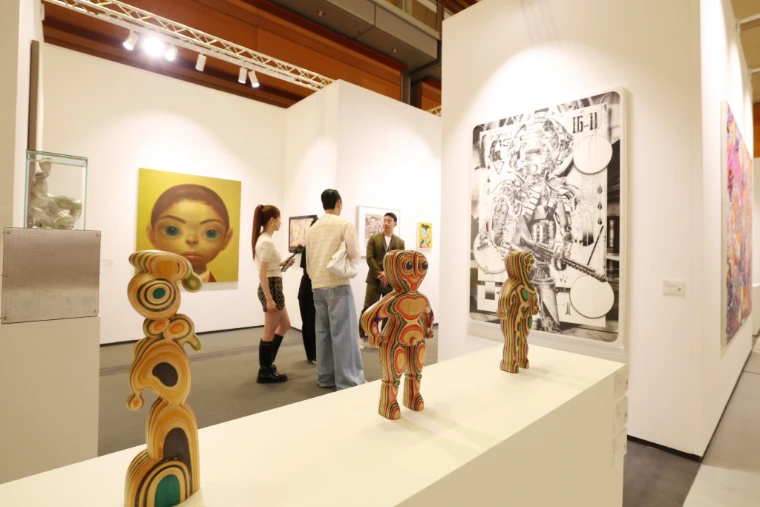 Visitors talk at a gallery booth hung with paintings, with three carved, multicoloured wooden figures on a white plinth in the foreground (2025).