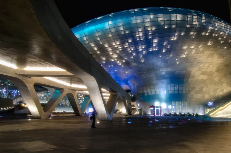 The curving, panelled shell of the Dongdaemun Design Plaza lit blue and white at night, seen from beneath a sweeping concrete walkway (2014).