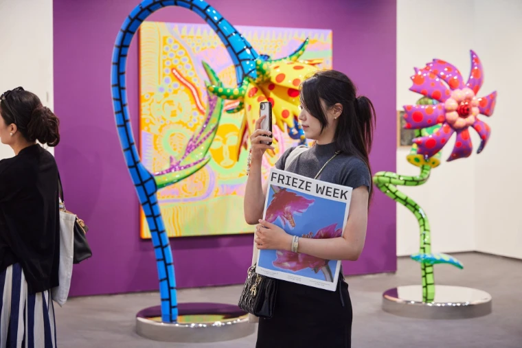 A woman holding a copy of Frieze Week photographs a painting and tall polka-dotted flower sculptures at a booth with a purple wall (2025).