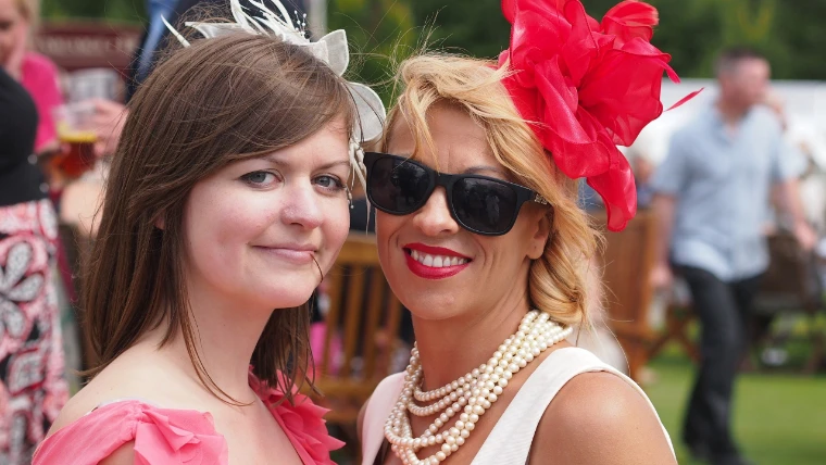 Two smiling women in summer dresses, one with a large red feather fascinator, sunglasses and pearls and the other a cream one, pose at the races (2012).