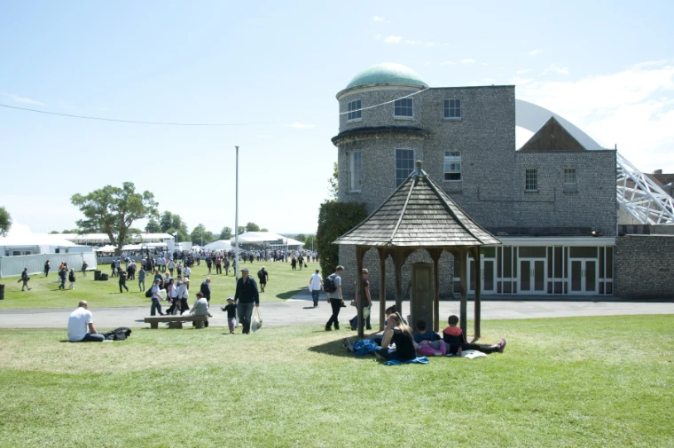 Visitors walk and sit on sunlit lawns beside a wooden gazebo and a domed flint building, with marquees in the distance (2014).