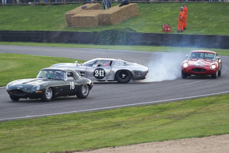 Two Jaguar E-types and a silver Bizzarrini race through a bend, one locking a wheel in tyre smoke, with marshals in orange on the bank behind (2014).