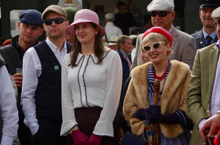 Spectators in period dress, among them a woman in a pink cloche hat and a woman in cat-eye sunglasses and a fur stole, stand watching (2019).