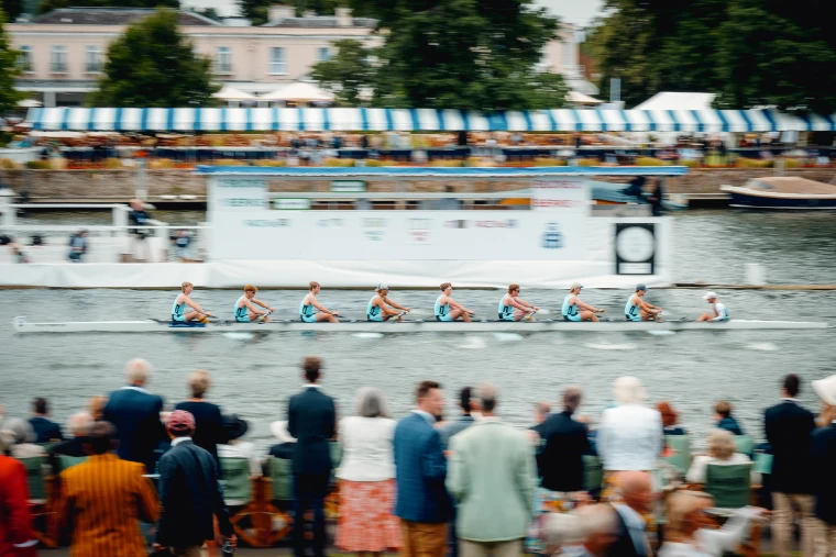 The Edinburgh University eight races past the grandstand, spectators in blazers watching from the bank.