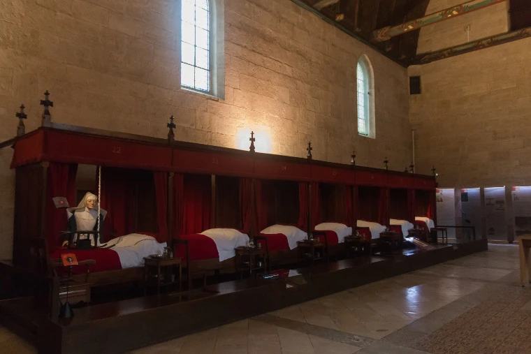 A row of curtained beds with red covers and white linen along the wall of the Room of the Poor in the Hôtel-Dieu, with a nun’s figure at the near end of the room (2016).