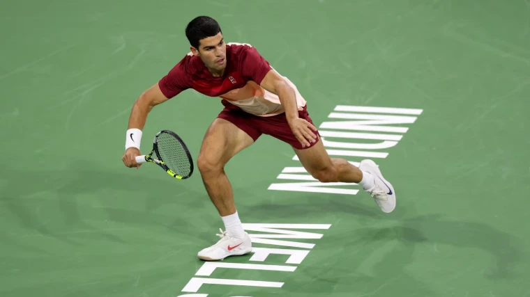 A player reaching across the green court under the lights at Indian Wells, California, the March fortnight of desert tennis in the season calendar of The Mansa Court