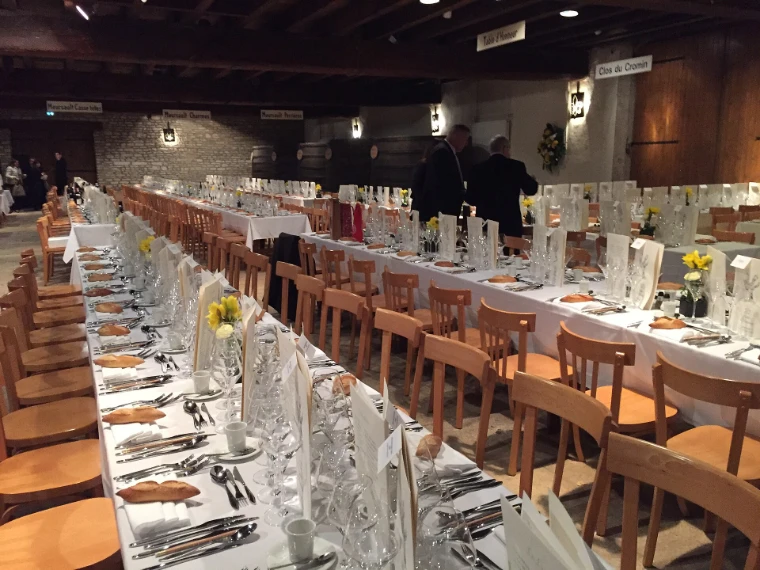 Long tables laid with white cloths, rows of glasses, bread and wooden chairs run the length of the beamed cuverie of the Château de Meursault before a Paulée lunch (2019).