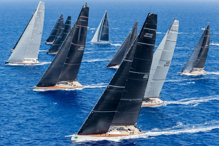A fleet of maxi yachts under dark and silver sails racing across open blue water, the class that contests Les Voiles de Saint-Barth, St. Barthélemy, a mid-April engagement in the season calendar of The Mansa Court