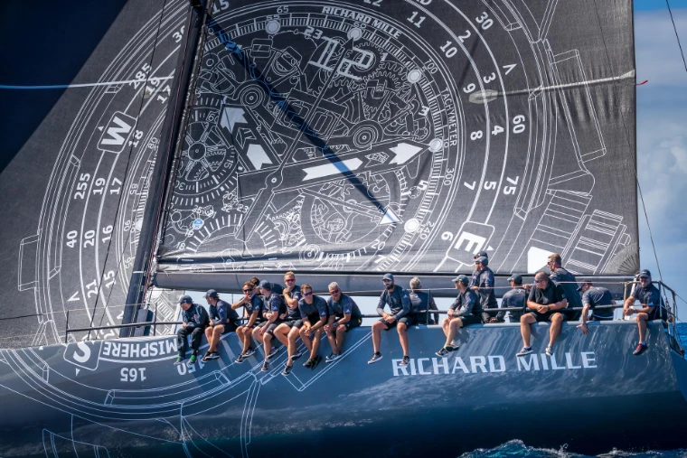 The crew of a maxi yacht lined along the rail beneath a mainsail printed with a watch dial at Les Voiles de Saint-Barth, St. Barthélemy, a mid-April engagement in the season calendar of The Mansa Court