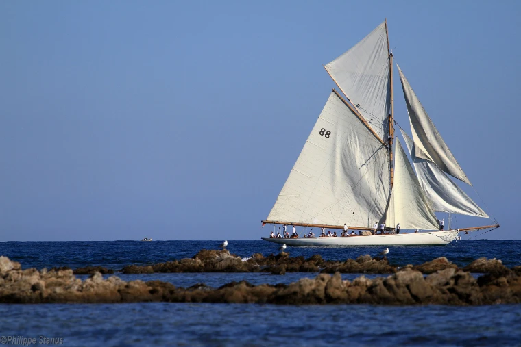 The classic gaff cutter Moonbeam III sails under a full cloud of white canvas past a low rocky reef, her crew lined along the deck (2014).