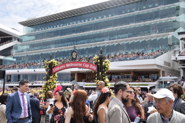 Racegoers in suits, dresses and fascinators beneath the rose-covered Melbourne Cup Day arch, with the grandstand behind (2015).