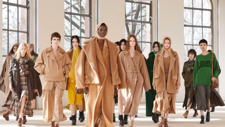 Models in camel coats, shearling and hooded knits walking through a daylit hall of tall arched windows at Milan Fashion Week, an engagement in the season calendar of The Mansa Court