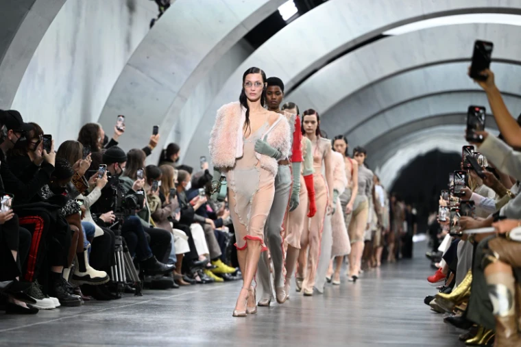 Models in blush chiffon and shearling walking a runway through a vaulted concrete hall as the front row films at Milan Fashion Week, an engagement in the season calendar of The Mansa Court